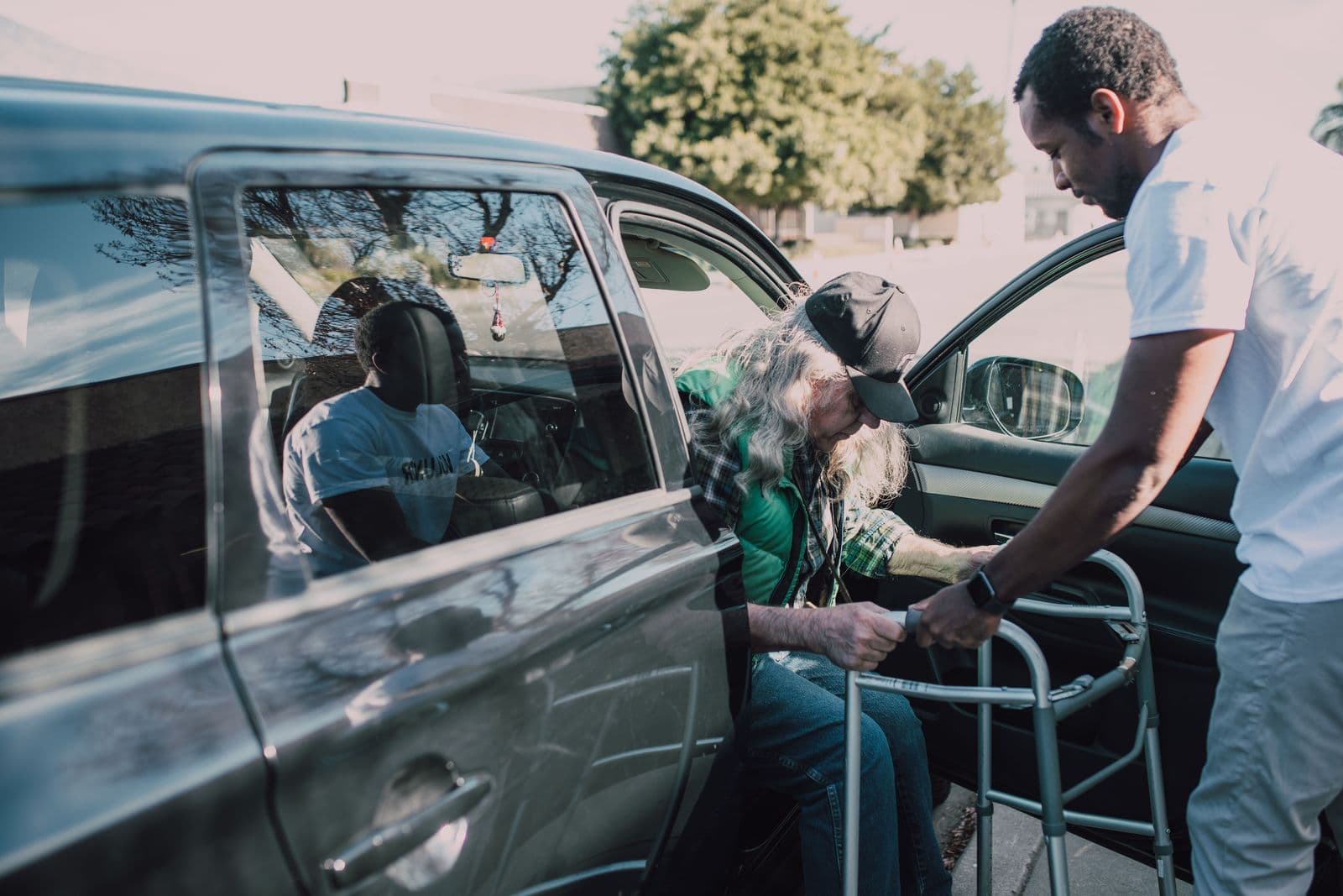 A driver steadying a walking frame as an older rider steps down from a vehicle at the kerbside.