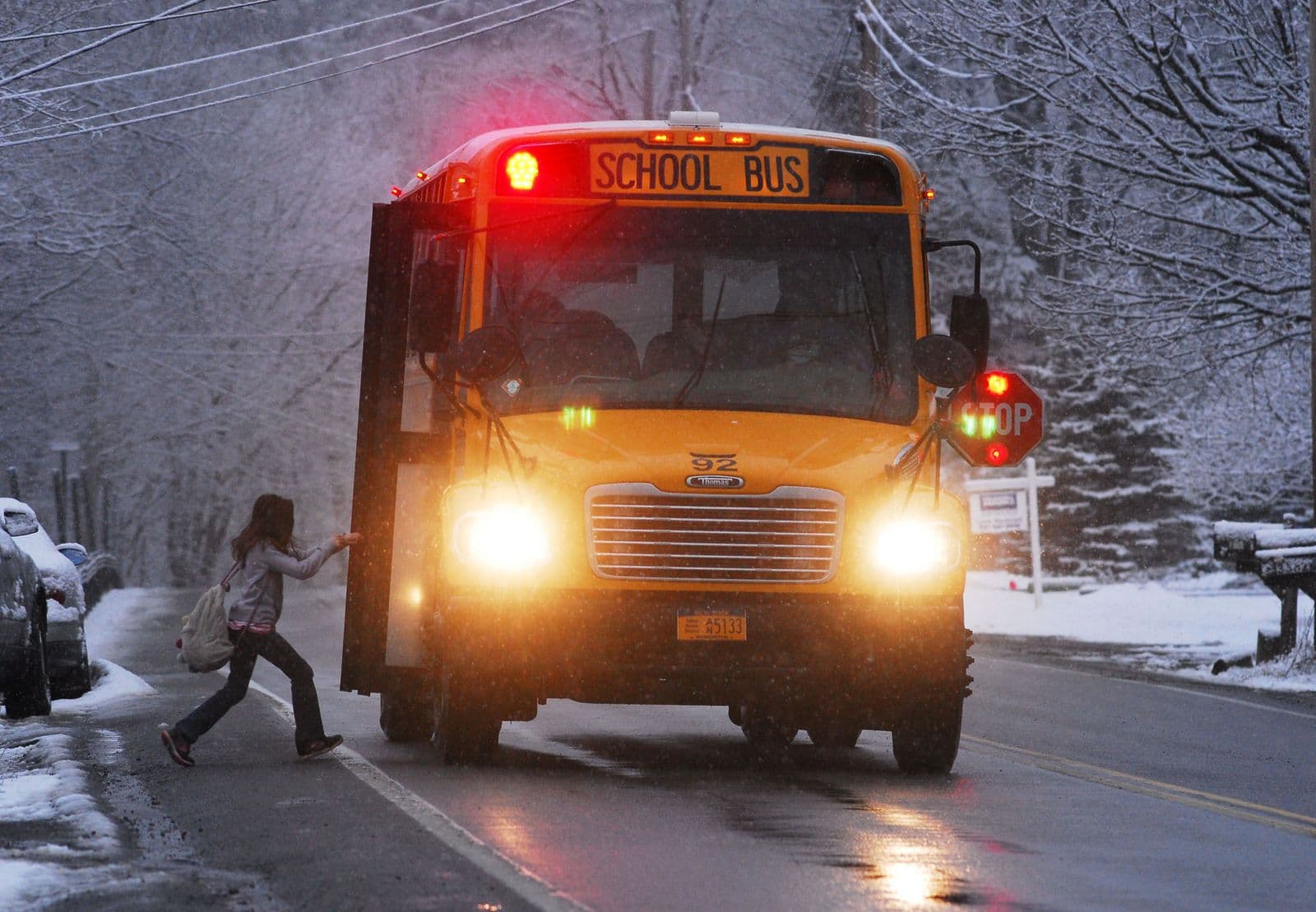 A yellow school bus stopped on a snowy road with its stop arm out and a child crossing in front