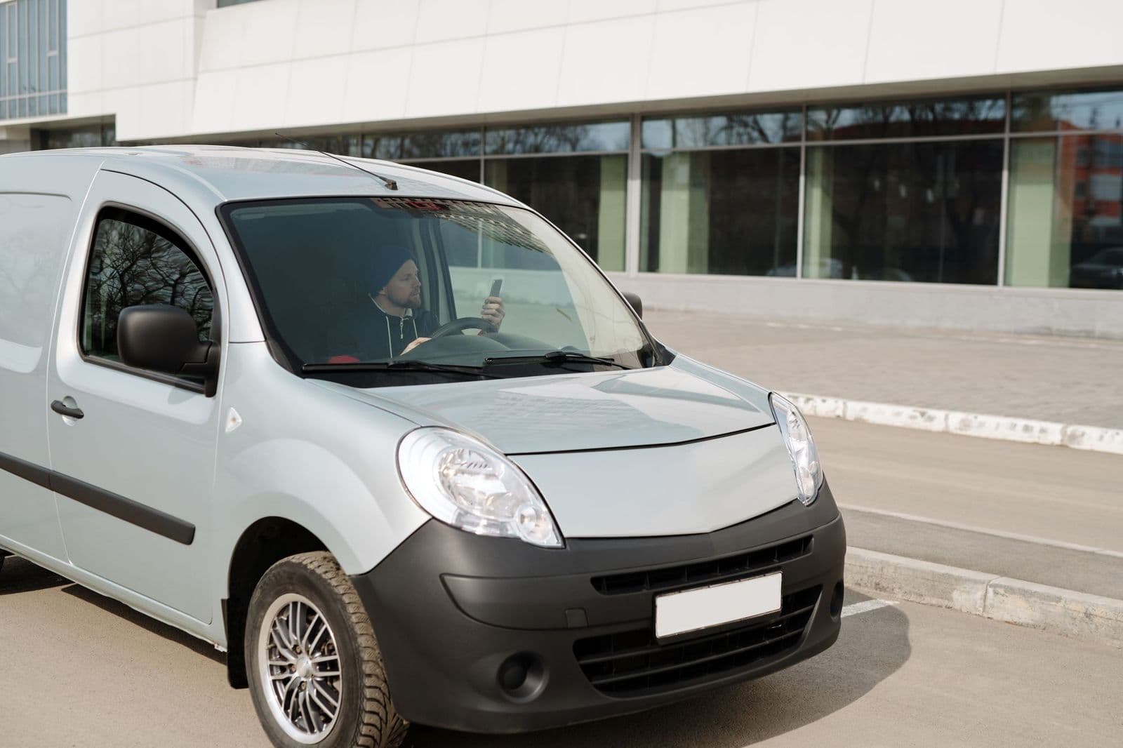 A driver sitting in a parked fleet van outside a facility, checking a phone before setting off.